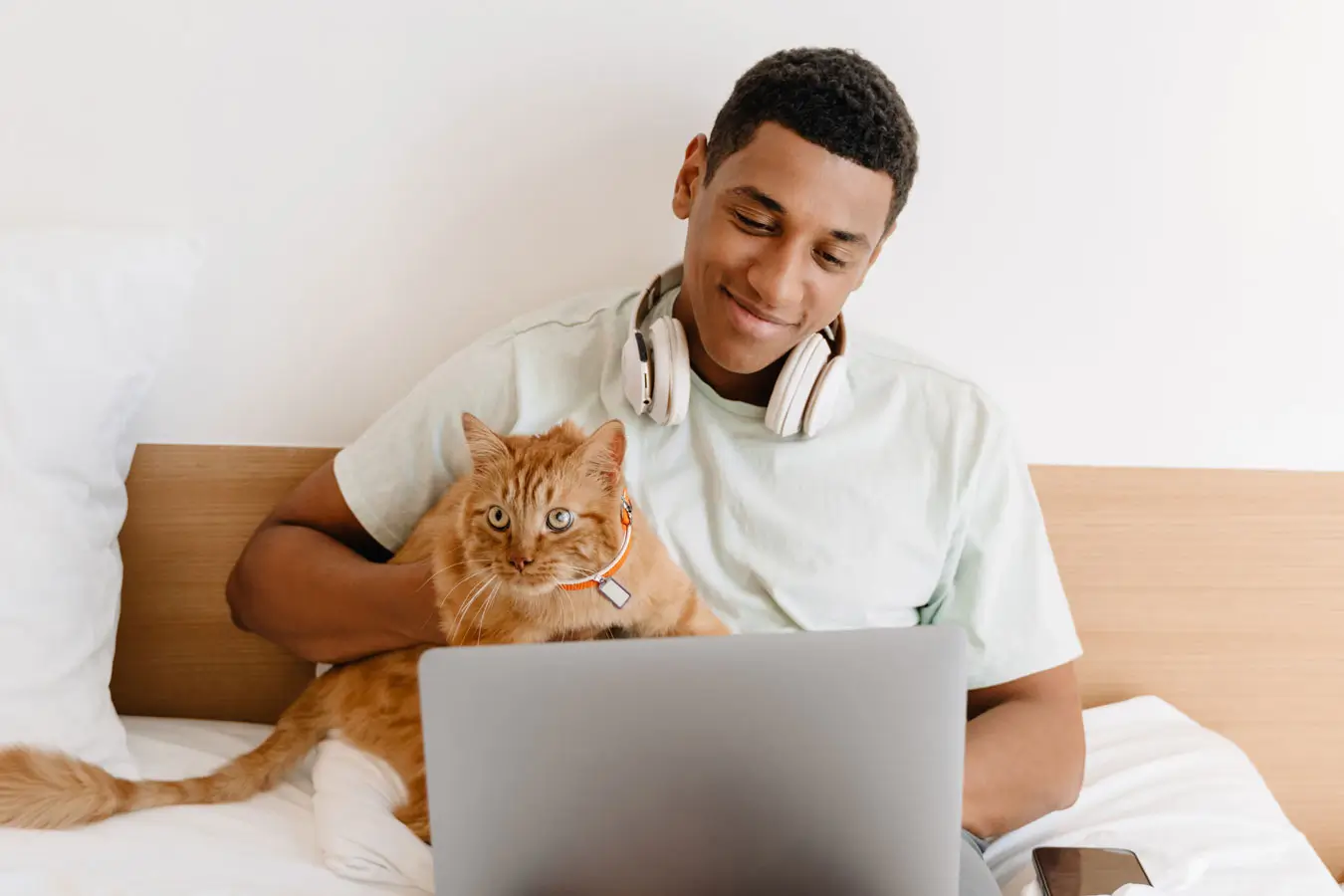 A man with headphones relaxes with his laptop on his lap. He holds a cute orange cat in his right arm.