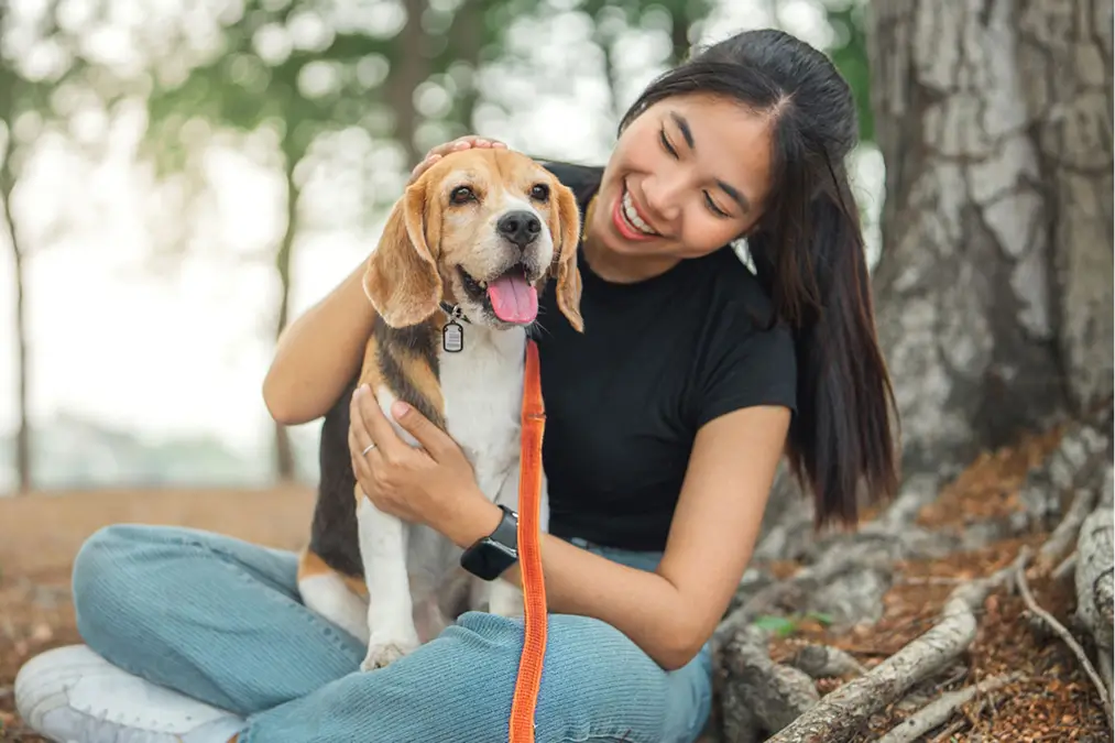 A young woman sits outside by a tree, holding her beagle.