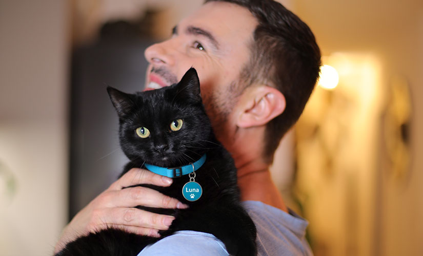 A man holds a black cat over his shoulder indoors.