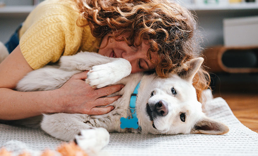 A woman is hugging a white dog wearing a blue collar and ID tag while lying on a rug indoors.