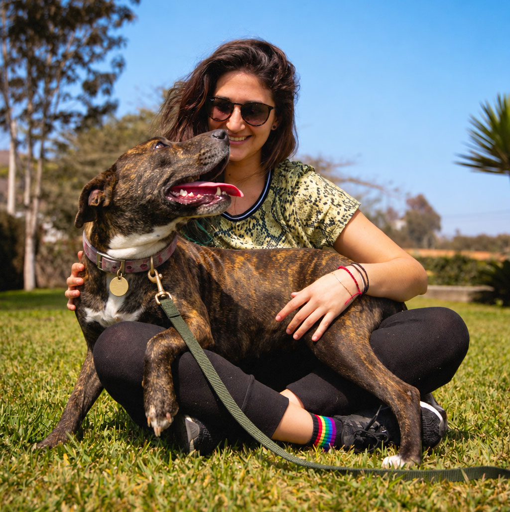 Woman sits on the grass cuddling a brindle dog wearing a collar and leash.