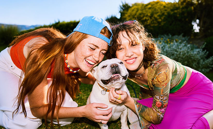 Two women cuddle a white English Bulldog outdoors in a grassy field with mountains in the background.