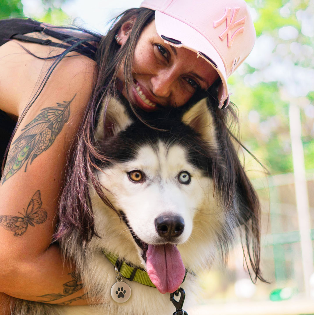 A woman hugs a black-and-white Husky wearing a green collar and paw-print ID tag outdoors.
