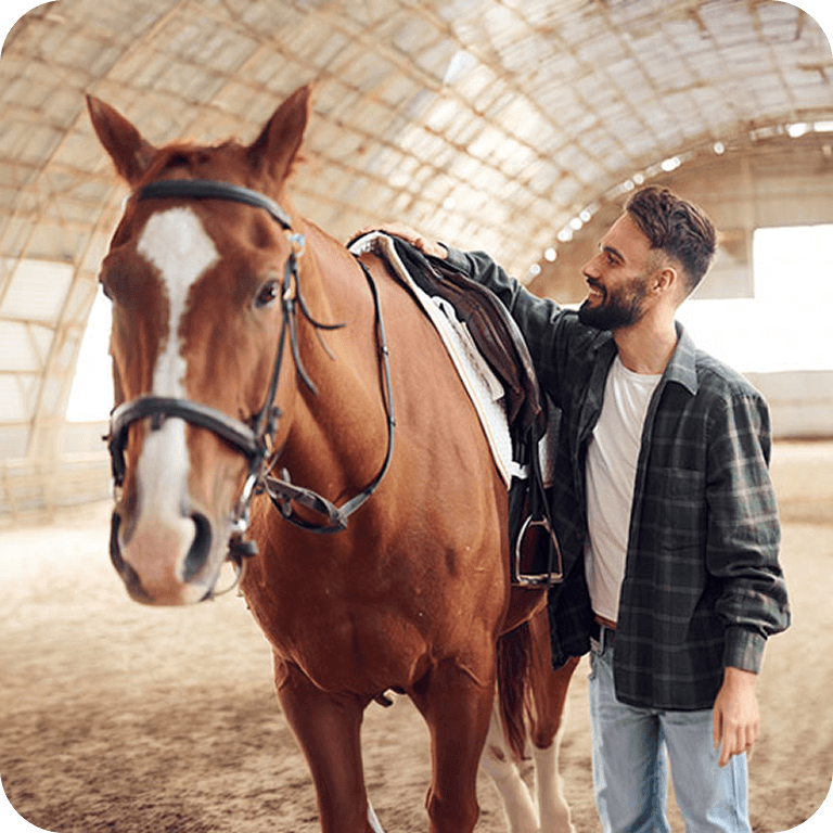 A smiling man stands beside a saddled horse in an indoor riding arena.