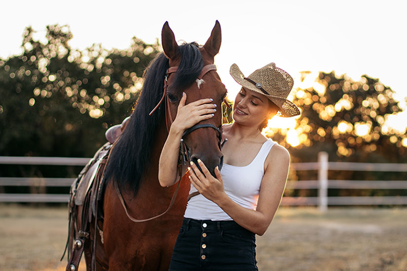 A woman in a cowgirl hat pets a brown horse in a riding arena at sunset.