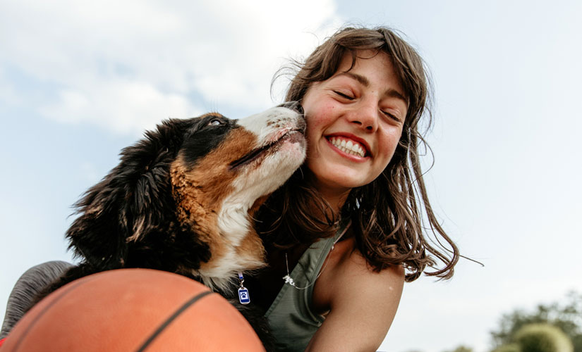 A smiling woman outdoors holding a basketball while a Bernese Mountain Dog affectionately licks their face, with trees and sky in the background.